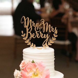 May include: A white wedding cake with a gold topper that says "Mr. & Mrs. Berry" with a leafy wreath around the text. The cake is decorated with white frosting and has a pink flower on the bottom tier.