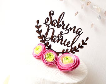 May include: A wooden cake topper with the names "Sabrina & Daniel" in elegant script, surrounded by a leafy wreath. Three pink and yellow flowers adorn the cake, which has a textured white surface. Decorative shells hang in the background.