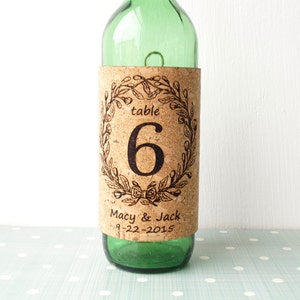 May include: Cork table number with a floral wreath design. The number 6 is displayed in the center. The text "table" is above the number. The text "Macy & Jack 9-22-2015" is below the number.