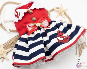 May include: A red and white striped sailor dress for a pet with a red anchor and a red bow. The dress has a white collar and a white lace trim. The dress is on a white background with a rope and starfish.