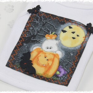 May include: White ribbed tank top with a Halloween-themed graphic. The graphic features a ghost wearing a pumpkin costume, a full moon, bats, and a spooky background. Black bat-shaped embellishments are sewn on the corners of the graphic.
