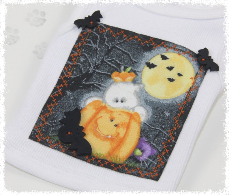 May include: White ribbed tank top with a Halloween-themed graphic. The graphic features a ghost wearing a pumpkin costume, a full moon, bats, and a spooky background. Black bat-shaped embellishments are sewn on the corners of the graphic.