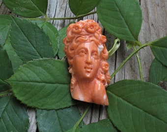 May include: A carved coral cameo of a woman's face with a floral design around her head. The cameo is a light orange color and is set against a background of green leaves.