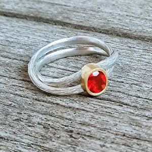 Könnte beinhalten: Ein silberner Ring mit einem gewebten Banddesign. Der Ring hat eine goldene Lünettenfassung, die einen runden, leuchtend roten Edelstein hält. Das Design des Rings ist modern und elegant, geeignet für den Alltag oder besondere Anlässe.