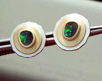 Puede incluir: Un par de pendientes de botón con una piedra central de ópalo oscuro ovalada, que muestra iridiscencia verde y azul. El ópalo está engastado en un bisel dorado, rodeado por un diseño de disco dorado y plateado. Los pendientes se muestran en una herramienta plateada.