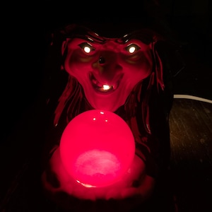 May include: A red, illuminated Halloween decoration featuring a witch's face with glowing white eyes and a toothy grin. The witch holds a glowing red orb. The background is dark, emphasizing the eerie glow of the decoration.