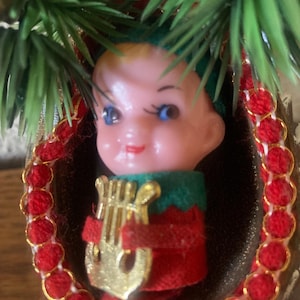 May include: A vintage-style Christmas ornament featuring a small plastic elf figurine with blue eyes and a pink face. The elf wears a green and red outfit and holds a gold-colored plastic lyre. It is nestled inside a brown walnut shell frame.