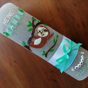 以下が含まれることがあります： グレーの洗礼用キャンドル。枝にぶら下がったナマケモノの置物、緑の葉、ターコイズブルーのリボン、そして「Meine Taufe」と「Marlo 22.01.2021」の文字が書かれている。