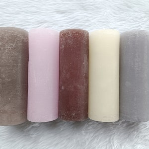 May include: Five pillar candles in different colors: brown, pink, brown, cream, and gray. The candles are arranged in a row on a white fuzzy surface.