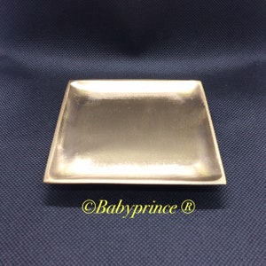 Könnte beinhalten: Ein quadratisches, goldfarbenes Tablett mit leicht erhöhtem Rand. Das Tablett besteht aus einem glatten, metallischen Material und hat einen dezenten Glanz.