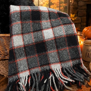 May include: A woven throw blanket featuring a plaid pattern in black, gray, and white with thin red accent lines. The blanket has a textured appearance and is finished with long, thick fringe along the bottom edge, displaying a classic check design.