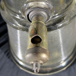 May include: Close-up of a glass bottle with a metal spout. The bottle has a textured surface and the spout has a silver finish.