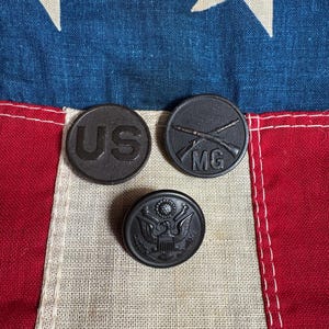 May include: Three dark gray military uniform buttons. One button has the letters "US", another has crossed rifles and "MG", and the third features an eagle emblem. The buttons are displayed on a vintage American flag.