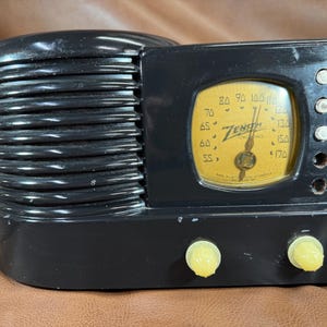 May include: A black Zenith radio with a horizontal ribbed speaker grille on the left side. The front features a yellow dial with black numbers and the Zenith logo. Two yellow knobs are positioned at the bottom, and four circular buttons are on the right.