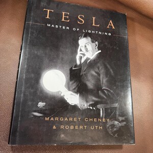May include: A hardcover book titled "TESLA MASTER OF LIGHTNING" featuring a black and white portrait of Nikola Tesla holding a glowing lightbulb. The book cover is dark with white lettering and the authors' names are visible.