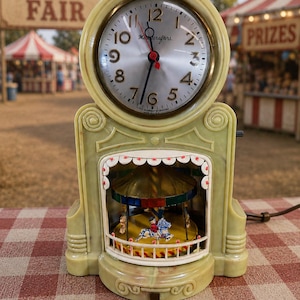 May include: A light green Mastercrafters clock with a round face, black numerals, and red hands. Below the clock face is a recessed area containing a miniature carousel with colorful horses and a striped canopy, enclosed by a white railing.