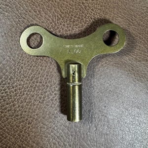 May include: A brass-colored key with a large, flat, T-shaped head and a round shaft. The head has two holes and the text "SWISS MADE 5.00" is printed on the top.