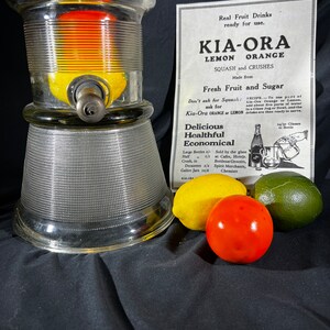 May include: A glass dispenser with a clear glass base and a textured glass top. The dispenser has a metal spout and is filled with lemons and oranges. The dispenser is sitting on a black surface with a yellow lemon, a red orange, and a green lime.