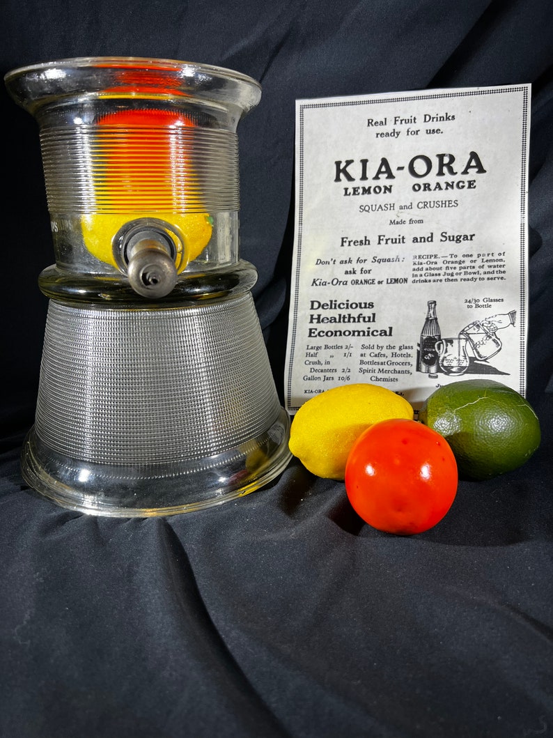 May include: A glass dispenser with a clear glass base and a textured glass top. The dispenser has a metal spout and is filled with lemons and oranges. The dispenser is sitting on a black surface with a yellow lemon, a red orange, and a green lime.