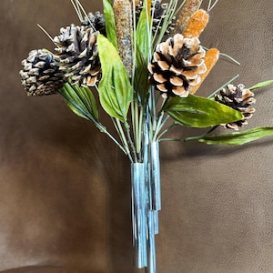 May include: A silver metal vase with a round base and multiple tubes for holding stems. The vase is filled with a bouquet of artificial pine cones and cattail-like stems.