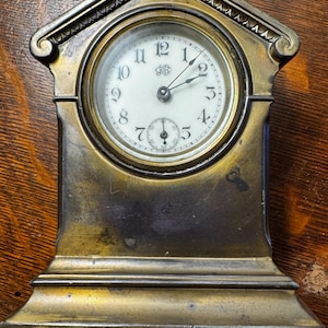 May include: A brass clock with a white face and black hands. The clock is shaped like a house and has a decorative design around the top. The clock face has the number 12 at the top and the numbers 1 through 11 going clockwise. The clock hands are pointing to 12:00.