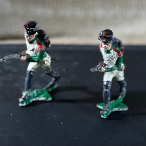 May include: Two toy soldiers in white and green uniforms with black hats and guns. The soldiers are standing on green bases.