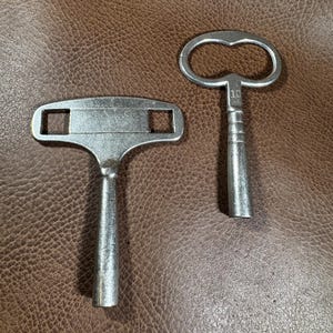 May include: Two silver-toned keys for winding a clock. The key on the left has a T-shaped handle and the key on the right has a rounded handle with the number "10" engraved on it.