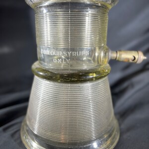 May include: A clear glass syrup dispenser with a ribbed design and a spout on the side. The dispenser has the text "For Our Syrups Only" etched on the side.