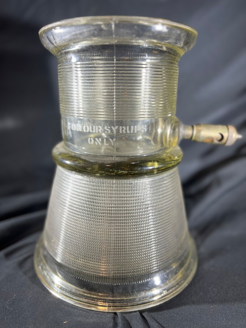 May include: A clear glass syrup dispenser with a ribbed design and a spout on the side. The dispenser has the text "For Our Syrups Only" etched on the side.