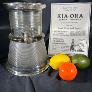 May include: A vintage glass citrus juicer with a silver-colored base and a clear glass top. The juicer has a spout for pouring juice and a handle for squeezing citrus fruits. The juicer is sitting on a black surface with three fruits: a yellow lemon, a red tomato, and a green lime.