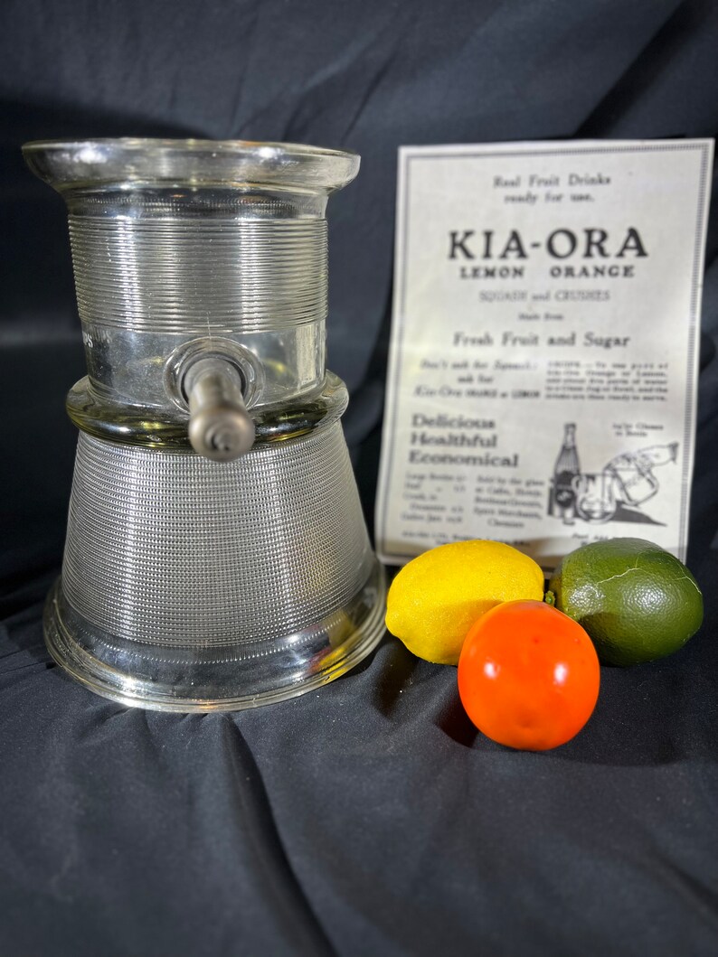 May include: A vintage glass citrus juicer with a silver-colored base and a clear glass top. The juicer has a spout for pouring juice and a handle for squeezing citrus fruits. The juicer is sitting on a black surface with three fruits: a yellow lemon, a red tomato, and a green lime.