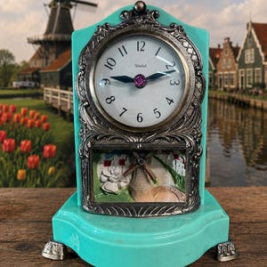May include: A turquoise mantel clock with a white face, black numerals, and the word United printed on the dial. The clock features an ornate silver-toned metal frame and a lower panel depicting a relief scene of a windmill and a figure.