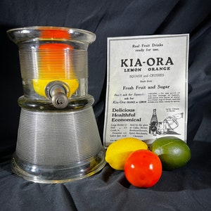 May include: A glass citrus juicer with a metal spout and a base with a textured pattern. The juicer is filled with a lemon and orange. The image also includes a vintage advertisement for Kia-Ora Lemon and Orange Squash and Crushes, made from fresh fruit and sugar. The ad includes a recipe for making the drink and information about where to buy it.