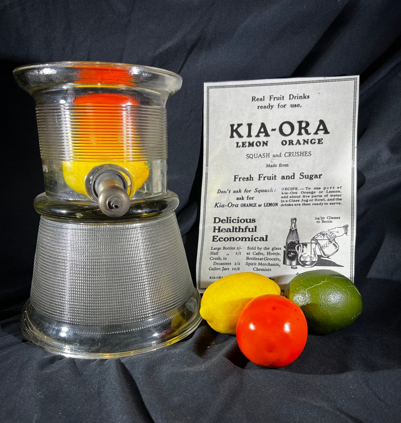 May include: A glass citrus juicer with a metal spout and a base with a textured pattern. The juicer is filled with a lemon and orange. The image also includes a vintage advertisement for Kia-Ora Lemon and Orange Squash and Crushes, made from fresh fruit and sugar. The ad includes a recipe for making the drink and information about where to buy it.