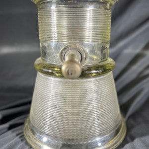 May include: A clear glass bottle with a textured surface and a metal spout. The bottle has a wide base and a narrow neck. The bottle is designed for dispensing liquids.