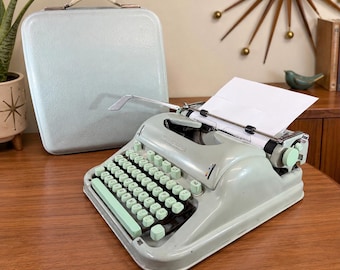 May include: A light mint green portable typewriter with a matching hard-shell carrying case. The typewriter features a QWERTY keyboard with round mint green keys, a metal carriage, and a sheet of white paper inserted into the platen roller.