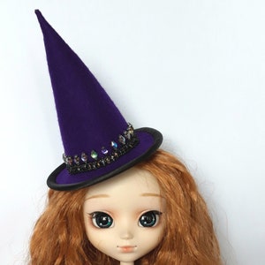 May include: A purple felt witch hat with a black brim and iridescent gem embellishments. The hat is worn by a doll with long, curly red hair and blue eyes.
