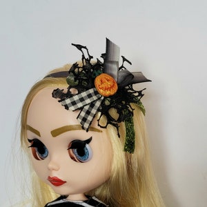 Op de afbeelding: Een zwart-wit geblokte strik met zwarte kant en een oranje pompoenknop, bevestigd aan een zwart hoofdband. Het hoofdband wordt gedragen door een pop met blond haar en blauwe ogen.