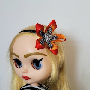 Op de afbeelding: Een pop met blond haar die een zwart hoofdband draagt met een rood en oranje strikje met een zwart-wit geruit midden.