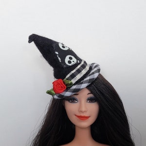 Op de afbeelding: Een zwart-witte heksenhoed met een schedelontwerp en een rode roosaccent. De hoed wordt gedragen door een pop met lang zwart haar.