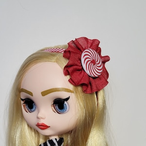 Op de afbeelding: Een pop met blond haar die een rood-wit snoepstokje hoofdband draagt met een rode strik.
