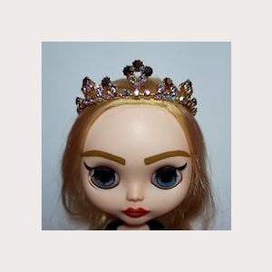 Peut inclure: Une poupée aux cheveux blonds portant une tiare dorée avec des pierres précieuses irisées et rouges.
