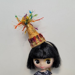 Puede incluir: Una muñeca con cabello negro que lleva un gorro de fiesta dorado con serpentinas de colores. El gorro tiene un borde dorado y rojo.