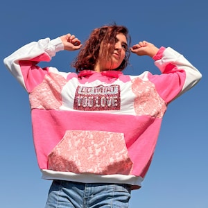 Pode incluir: Uma camisola com capuz e mangas compridas com um design de patchwork em branco e tons de rosa. A frente apresenta um emblema retangular com o texto DO WHAT YOU LOVE em letras brilhantes. A peça inclui painéis com textura de renda.
