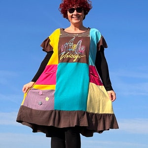 May include: A woman wearing a colorful patchwork dress with a Chicago skyline graphic. The dress has a brown, yellow, pink, turquoise, and purple patchwork design. The woman is wearing sunglasses and has red hair.