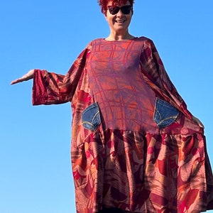 Op de afbeelding: Een vrouw met rood haar draagt een lange, vloeiende jurk met een abstract rood, oranje en bruin print. De jurk heeft twee grote denim zakken aan de zijkanten. Ze staat voor een blauwe lucht.