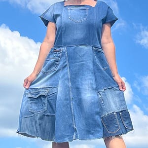 May include: A denim dress with a square neckline and short sleeves. The dress features multiple pockets and patchwork details, showcasing various shades of blue denim. The model is wearing sunglasses and smiling against a blue sky.