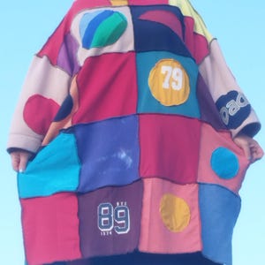 Op de afbeelding: Een kleurrijke patchwork tuniek met rode, blauwe, roze en gele vierkanten. De tuniek heeft de nummers "79" en "89" op sommige van de patches. De mouwen zijn lang en de stijl is los en comfortabel.