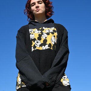 Pode incluir: Uma camisola com capuz preta com um painel central e detalhes laterais com uma estampa amarela e preta do Mickey Mouse. A peça inclui um tecido texturizado de malha waffle na frente e nas mangas, mangas compridas e um capuz.