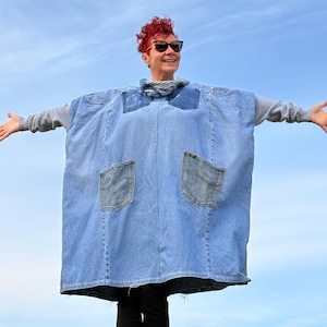 Pode incluir: Um poncho de ganga azul claro com dois bolsos traseiros e uma gola alta cinzenta. O poncho é usado sobre uma camisa cinzenta de manga comprida. A pessoa está a usar óculos de sol e tem cabelo ruivo.